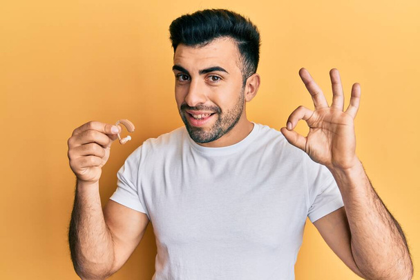 Homem hispânico jovem segurando fone de ouvido surdez fazendo sinal ok com os dedos, sorrindo amigável gesto excelente símbolo  - Foto, Imagem