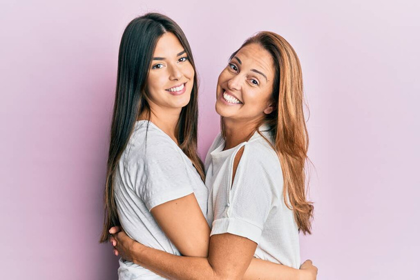 Beautiful hispanic mother and daughter smiling happy hugging over isolated pink background. - Foto, imagen