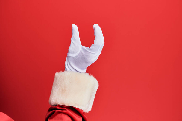 Hand eines Mannes im Weihnachtsmann-Kostüm und Handschuhen über rotem Hintergrund, der unsichtbare Dinge pflückt und nimmt und mit den Fingern Gegenstände hält, die Raum zeigen  - Foto, Bild