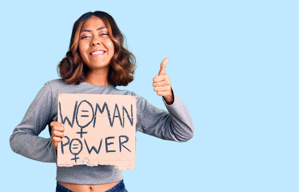 Young beautiful mixed race woman holding woman power banner smiling happy and positive, thumb up doing excellent and approval sign  - Photo, Image