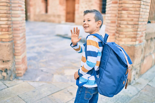 Adorable caucasian student boy smiling happy saying goodbye with hand at the school. - 写真・画像