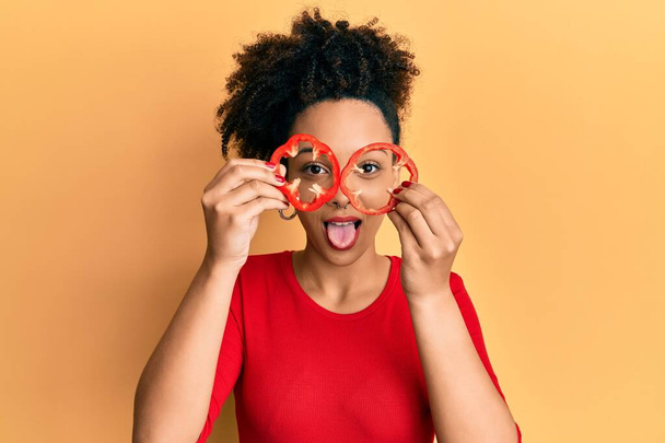 Young african american girl holding red pepper as a glasses sticking tongue out happy with funny expression.  - Photo, Image