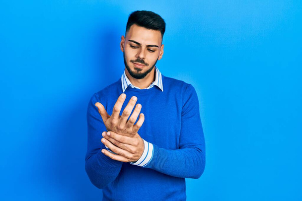 Junger hispanischer Mann mit Bart trägt lässigen blauen Pullover und leidet unter Schmerzen an Händen und Fingern, Arthritis-Entzündung  - Foto, Bild