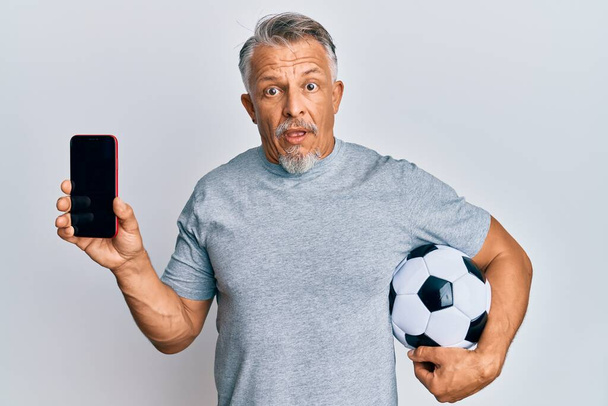 Middle age grey-haired man holding football ball showing smartphone clueless and confused expression. doubt concept.  - Foto, Bild