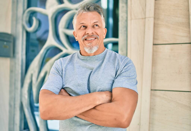 Hombre hispano de pelo gris de mediana edad con los brazos cruzados sonriendo feliz en la ciudad. - Foto, imagen