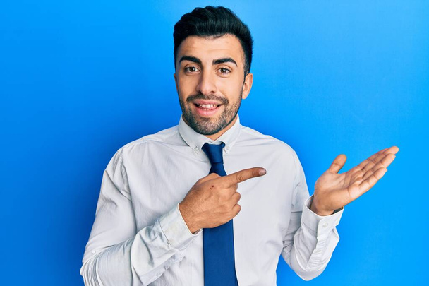 Jovem hispânico vestindo roupas de negócios espantado e sorrindo para a câmera ao apresentar com a mão e apontando com o dedo.  - Foto, Imagem