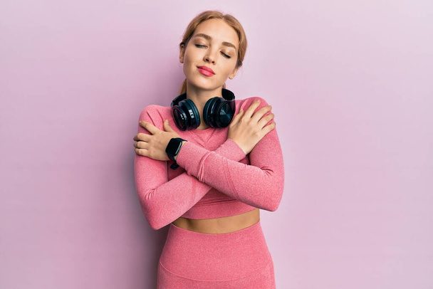 Beautiful blonde caucasian woman wearing gym clothes and using headphones hugging oneself happy and positive, smiling confident. self love and self care  - Photo, Image