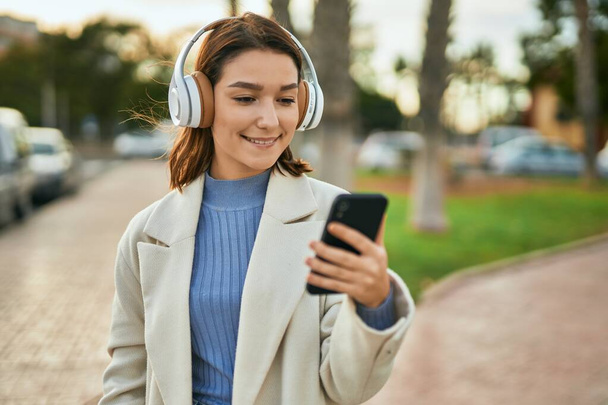 Young hispanic woman smiling happy using smartphone and headphones at the city. - Фото, изображение