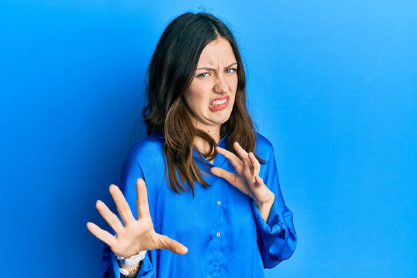 Νεαρή μελαχρινή γυναίκα φορώντας casual μπλε πουκάμισο αηδιασμένη έκφραση, δυσαρεστημένη και φοβισμένη να κάνει το πρόσωπο αηδίας επειδή αντίδραση αποστροφής. με τα χέρια σηκωμένα  - Φωτογραφία, εικόνα
