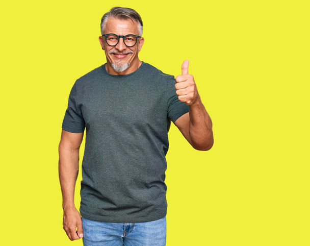 Grauhaariger Mann mittleren Alters in lässiger Kleidung und Brille, der glücklich und positiv lächelt, Daumen hoch und Zeichen der Anerkennung  - Foto, Bild