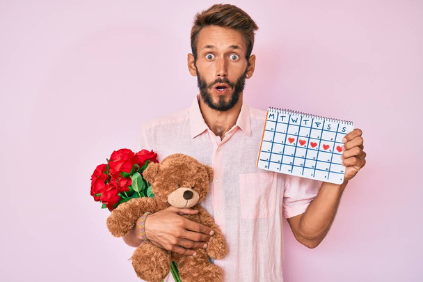 Handsome caucasian man with beard holding bear and bouquet of flowers for anniversary afraid and shocked with surprise and amazed expression, fear and excited face.  - Фото, зображення
