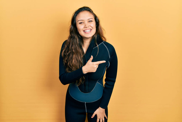 Young hispanic girl wearing diver neoprene uniform cheerful with a smile on face pointing with hand and finger up to the side with happy and natural expression  - Fotografie, Obrázek