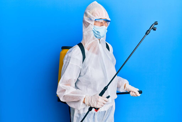 Covid specialist draagt decontaminatie beschermende pak en ontsmettingsmiddel spray, ontsmetten en reinigen oppervlak, staande over blauwe achtergrond - Foto, afbeelding