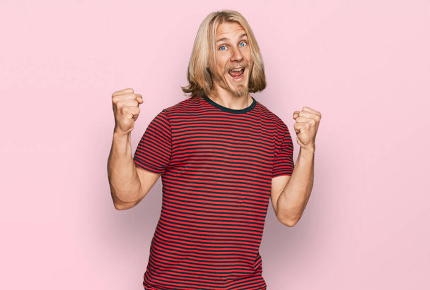 Caucasian man with blond long hair wearing casual striped t shirt celebrating surprised and amazed for success with arms raised and open eyes. winner concept.  - Photo, Image