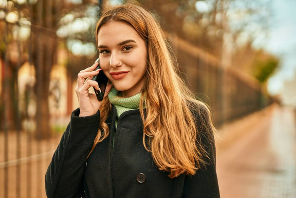 Chica rubia joven sonriendo feliz hablando en el teléfono inteligente en la ciudad. - Foto, Imagen