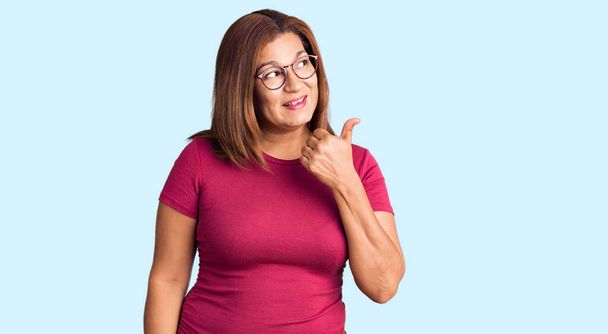 Middle age latin woman wearing casual clothes and glasses smiling with happy face looking and pointing to the side with thumb up.  - Фото, зображення