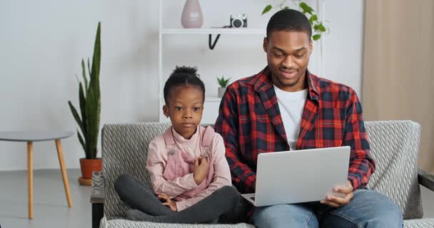 Afro americano dos personas, padre e hija sentado en sofá hombre mira portátil juega juego en línea funciona largo tiempo mientras niño lindo niña habla explica expresar dudas extiende sus brazos a los lados - Metraje, vídeo