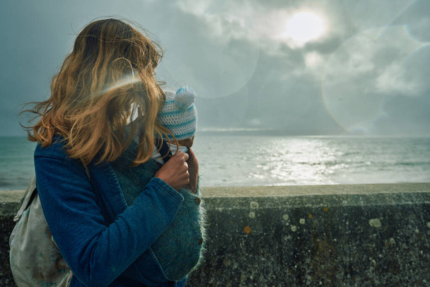 A young mother with her baby in a sling is standing by the sea and looking over a wall - Fotoğraf, Görsel