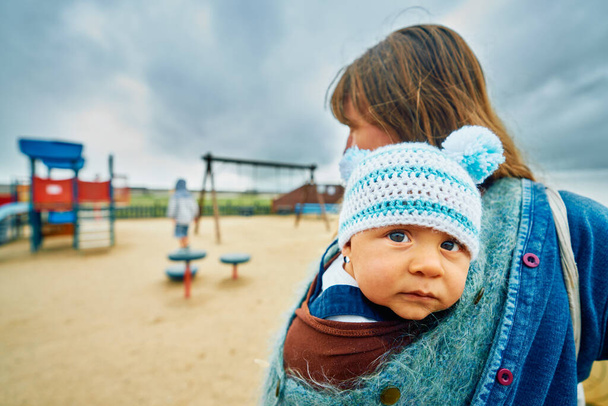 A young mother is standing in a playground with her baby in a sling - Photo, image