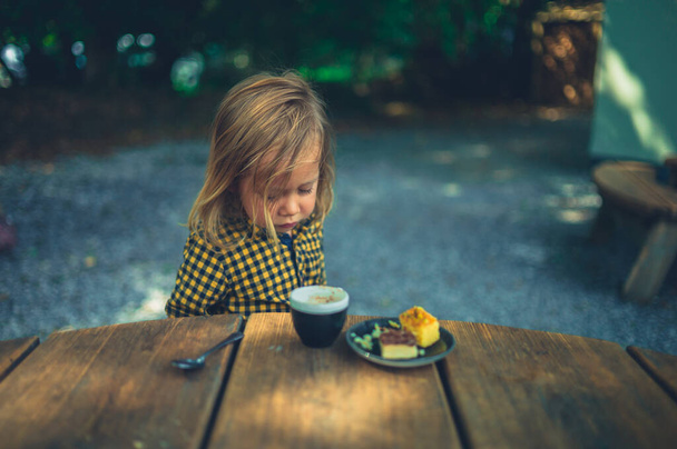 A little preschooler is drinking coffee and eating cake outdoors at a cafe table - Fotografie, Obrázek