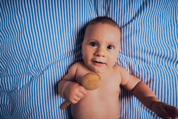 A little baby is lying on a bed with a wooden rattle - Foto, Bild