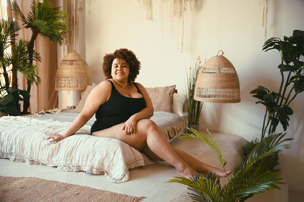 Mooie ronde oversized Afrikaanse zwarte vrouw afro haar liggend op bed eco vriendelijke slaapkamer gezellig interieur met home planten. Lichaamsimperfectie, lichaamsacceptatie, lichaamspositief en diversiteitsconcept  - Foto, afbeelding