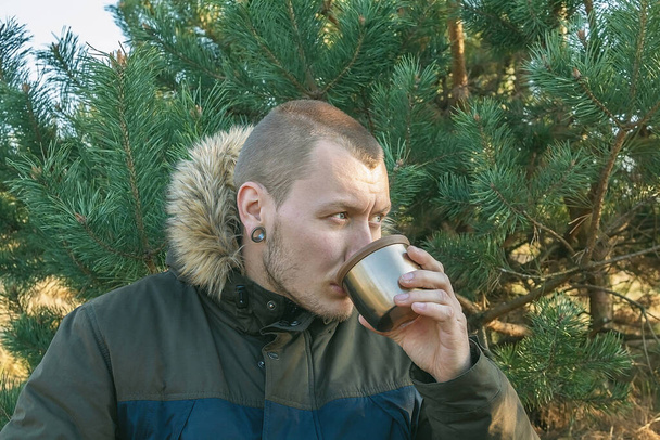 Nuori mies juo kuumaa teetä luonnossa olevasta mukista. Lepää ennen matkaa - Valokuva, kuva