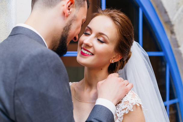 Braut und Bräutigam stehen vor blauen Fenstern und lächeln. Der Bräutigam umarmt die Braut mit der Hand am Hals . - Foto, Bild