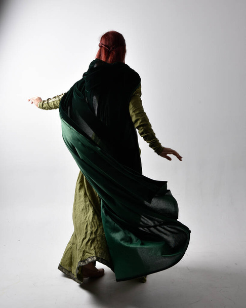 full length portrait of red haired girl wearing celtic, green medieval gown with shadowy backlighting. Standing pose isolated against a studio background. - Photo, Image