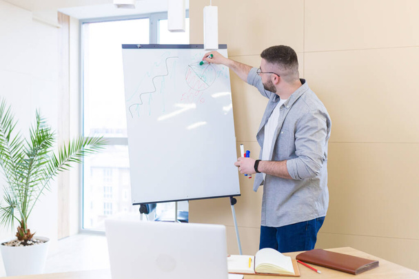 young business man in casual clothes online meeting presentation or training using a laptop webcam and a flipchart with markers. Businessman or coach teaches at home explaining a whiteboard chart - Fotoğraf, Görsel