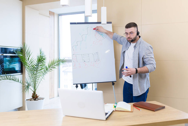 young business man in casual clothes wearing a headset online meeting presentation or training using a laptop webcam and a flipchart. Businessman or coach teaches at home explaining a whiteboard chart - Foto, immagini