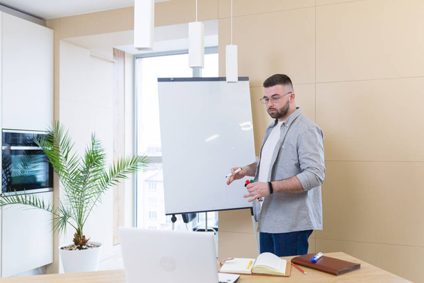 young business man in casual clothes online meeting presentation or training using a laptop webcam and a flipchart with markers. Businessman or coach teaches at home explaining a whiteboard chart - Φωτογραφία, εικόνα