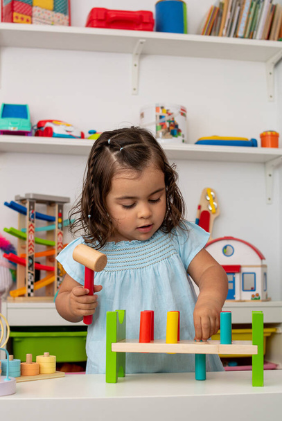 Two-year-old girl of Caucasian origin, playing with a hammer and some pieces of wood that she hits, in the nursery. - Photo, Image