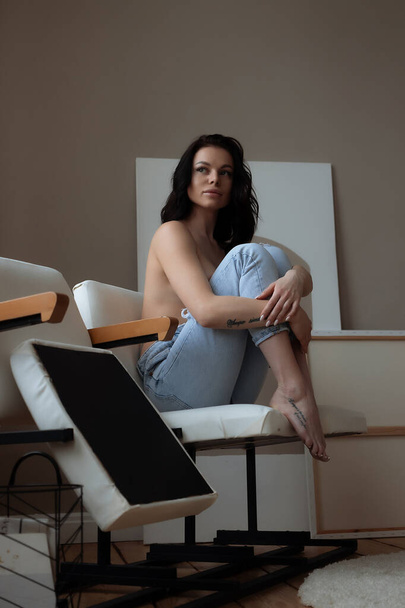 Sensual joven hembra en jeans cubriendo el pecho desnudo con la mano mientras se relaja sola en silla en acogedora habitación en casa - Foto, Imagen