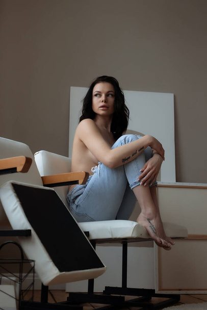 Sensuele jonge vrouw in jeans bedekken naakte borst met de hand, terwijl ontspannen alleen in stoel in gezellige kamer thuis - Foto, afbeelding