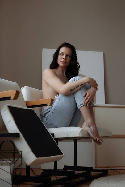 Чувственная молодая женщина в джинсах, прикрывающая обнаженную грудь рукой, расслабляясь в одиночестве в кресле в уютной комнате дома - Фото, изображение