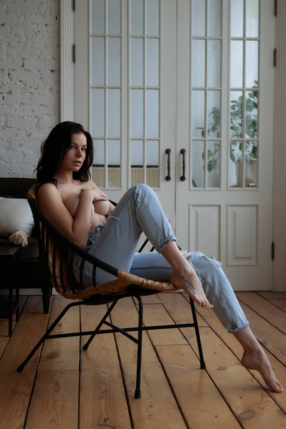 Sensual jovem mulher em jeans cobrindo peito nu com a mão enquanto relaxa sozinho na cadeira no quarto acolhedor em casa - Foto, Imagem