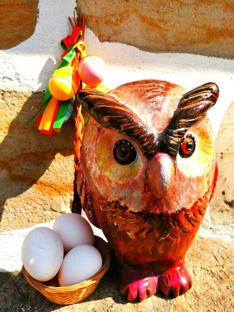 Пасхальная сова. Красочная статуэтка совы, выложенная маленькой корзиной с чистыми белыми куриными яйцами и небольшой плетеной плетеной плетью с цветными яйцами перед стеной из песчаника.  - Фото, изображение