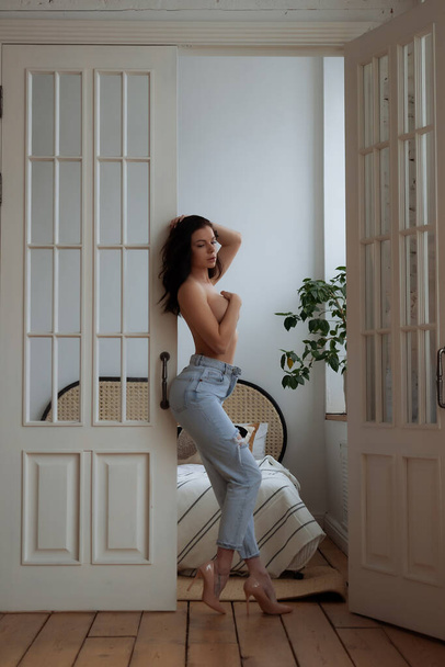 Full body of barefoot young slim female in trendy ripped jeans covering naked breast with hand while leaving bedroom at home - Photo, Image