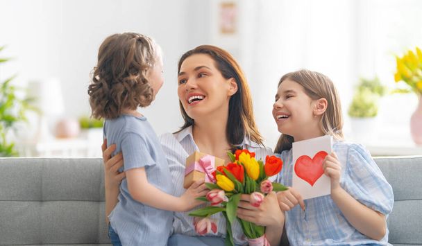 Happy mother's day! Children daughters are congratulating mom and giving her postcard and flowers. Mum and girls smiling and hugging. Family holiday and togetherness. - Fotó, kép