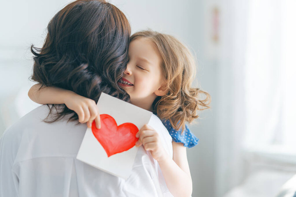 Happy mother's day! Child daughter is congratulating mom and giving her postcard. Mum and girl smiling and hugging. Family holiday and togetherness. - Φωτογραφία, εικόνα