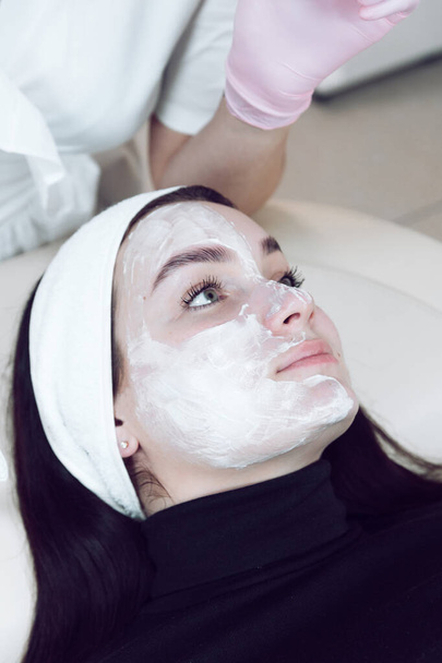 Egy nő arcplasztikai maszkkal. Kozmetológiai eljárás egy szépségszalonban. Arcápolás. Gyönyörű nő fehér kötéssel a fején és kozmetikai maszkkal a spa szalonban.. - Fotó, kép