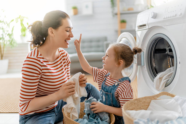 Beautiful young woman and child girl little helper are having fun and smiling while doing laundry at home. - Фото, изображение