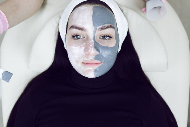 Een vrouw met een gezichtsmasker. Cosmetologische ingreep in een schoonheidssalon. Gezichtsverzorging. Mooie vrouw met een wit schoonheidsverband op haar hoofd en een cosmetisch gezichtsmasker in de spa salon. - Foto, afbeelding