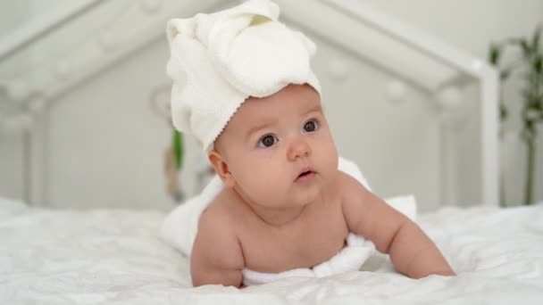 Ένα όμορφο νεογέννητο κοριτσάκι με μια πετσέτα στο κεφάλι ξαπλώνει σε ένα λευκό κρεβάτι, κοιτάζει τριγύρω και χαμογελάει. Έννοια ευτυχισμένων παιδιών. - Πλάνα, βίντεο