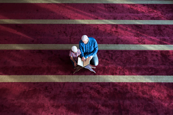 Мусульманский отец и сын молятся вместе. Мусульманский папа и сын молятся в мечети и читают священный Коран внутри мечети. - Фото, изображение