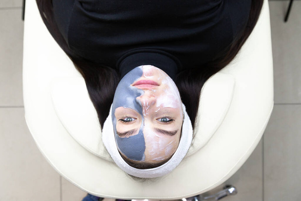 Een vrouw met een gezichtsmasker. Cosmetologische ingreep in een schoonheidssalon. Gezichtsverzorging. Mooie vrouw met een wit schoonheidsverband op haar hoofd en een cosmetisch gezichtsmasker in de spa salon. - Foto, afbeelding