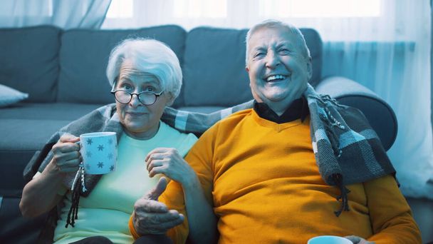 Heureux vieux couple riant regarder la télévision et boire une boisson chaude couverte de couverture - Photo, image