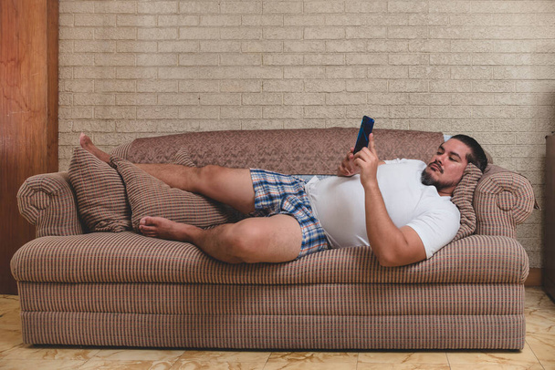 Μια τυπική πατάτα καναπέ - Ένας άνθρωπος με τα εσώρουχά του που βρίσκεται στον καναπέ και βλέποντας βίντεο, παίζοντας παιχνίδια για κινητά ή στα μέσα κοινωνικής δικτύωσης. - Φωτογραφία, εικόνα