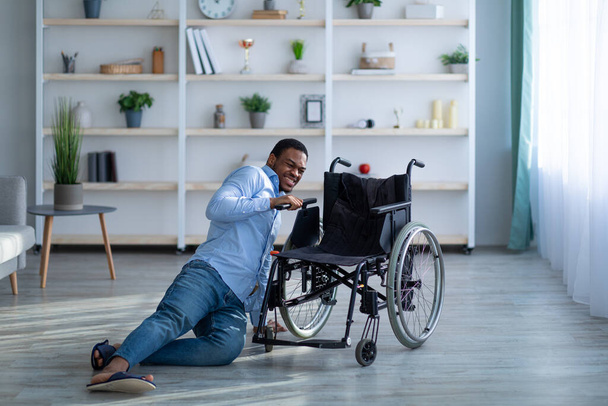 Giovane uomo nero con disabilità sdraiato sul pavimento dopo essere caduto dalla sedia a rotelle a casa - Foto, immagini
